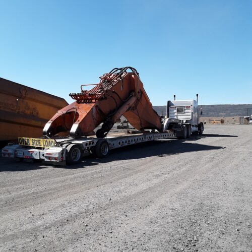 A large orange excavator arm is loaded on a flatbed truck, designated as an oversized load, in a gravel yard under a clear blue sky.
