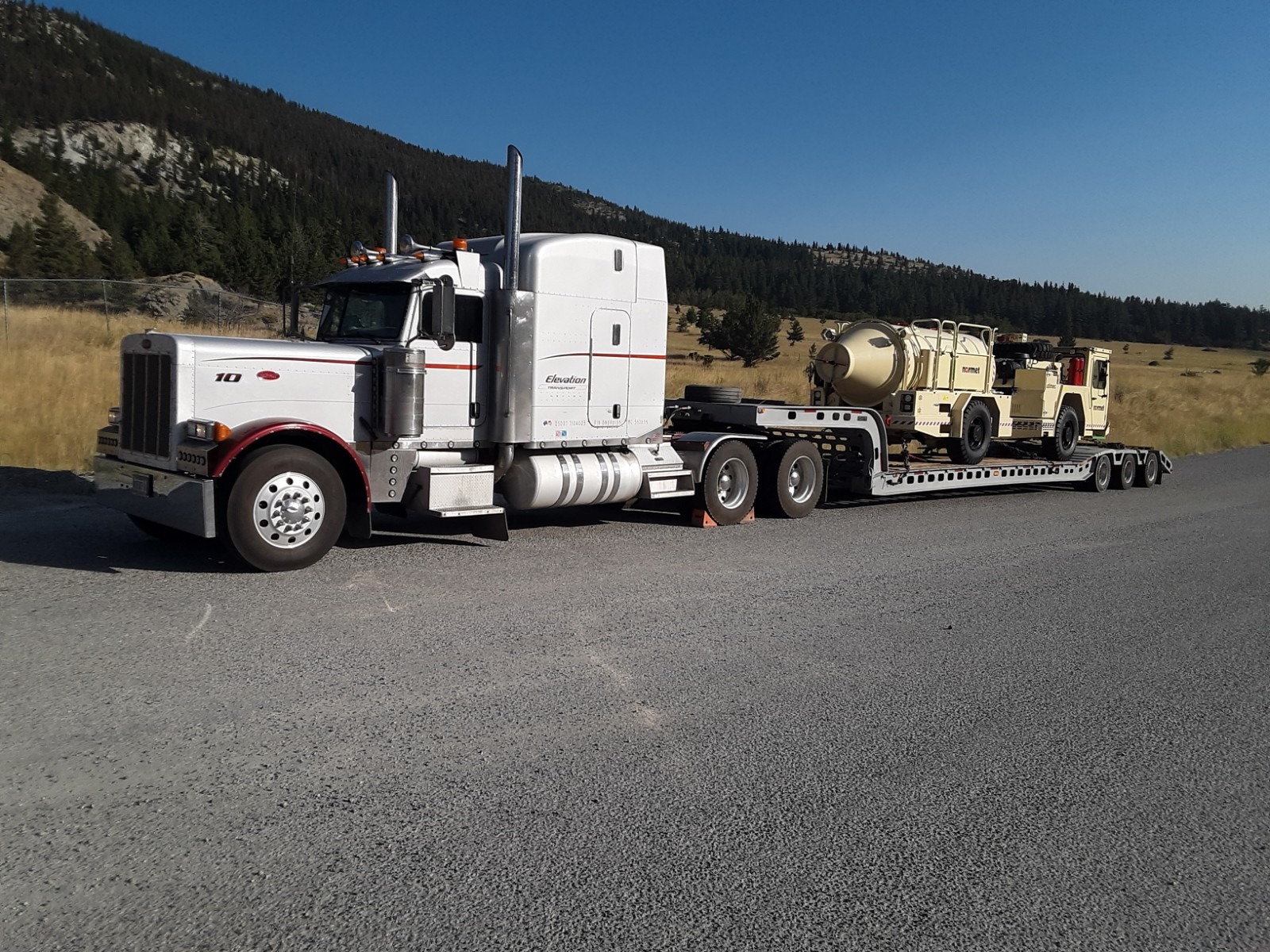 A white semi-truck with a red stripe hauls two military vehicles on a flatbed trailer, parked on a gravel road surrounded by mountains and grass.