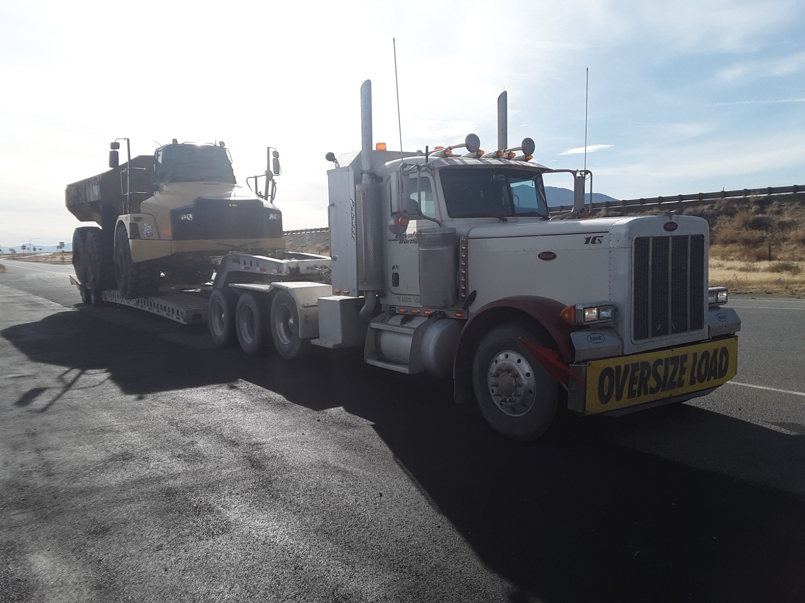 A tractor trailer transporting an oversized yellow dump truck on a flatbed trailer along a roadside. The scene highlights heavy freight transport.
