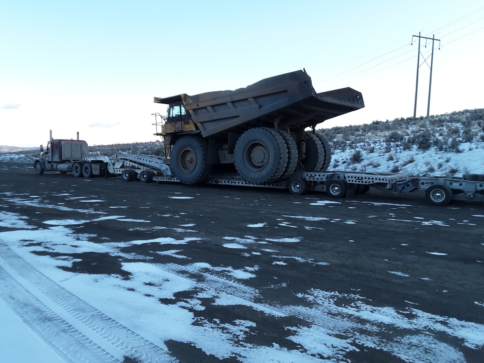 A large mining dump truck is loaded onto a flatbed trailer, parked on a snowy road with sparse vegetation and power lines in the background.