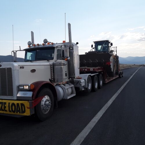 A large truck with a "Size Load" sign transports a heavy construction machine on a flatbed trailer, set against a scenic mountainous backdrop.