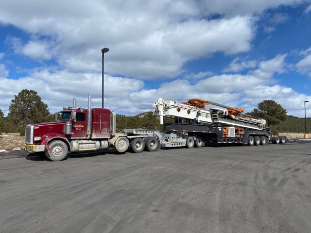 A red semi-truck with a long flatbed trailer transports a large piece of equipment against a backdrop of cloudy skies and trees.