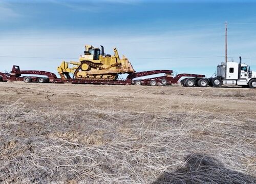 A large yellow bulldozer is loaded onto a flatbed truck on a barren landscape under a clear blue sky, showcasing heavy machinery transportation.
