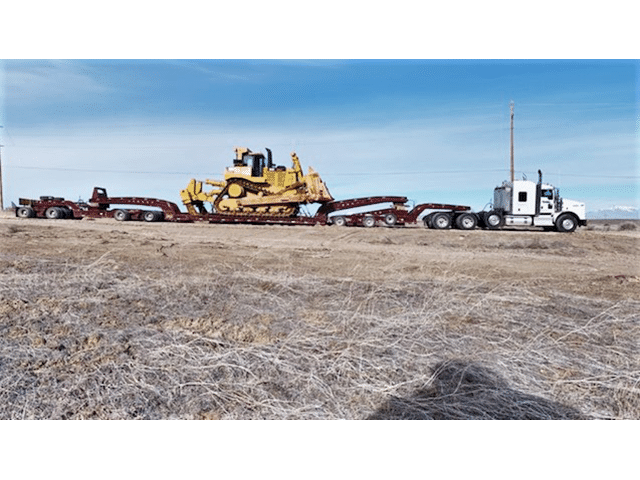 A large yellow bulldozer is loaded on a flatbed truck, transporting it across an open, dry landscape under a clear blue sky.