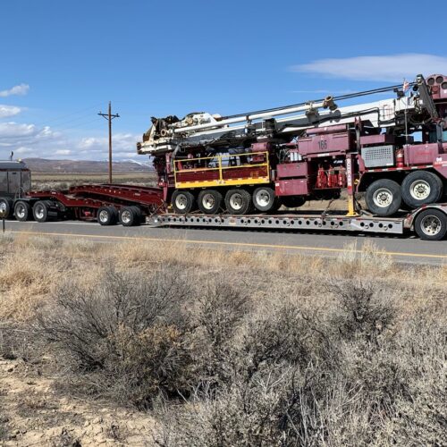 Large flatbed trailer transporting heavy machinery, likely an oil rig, on a rural road, with distant mountains and clear blue sky in the background.