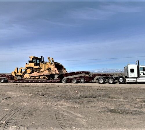 A large yellow bulldozer is loaded on a flatbed trailer being towed by a white truck on a gravel road against a clear sky.