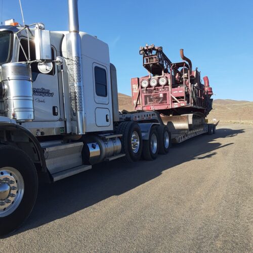 A large silver semi-truck transports heavy mining equipment on a flatbed trailer, set against a clear blue sky and mountainous terrain.