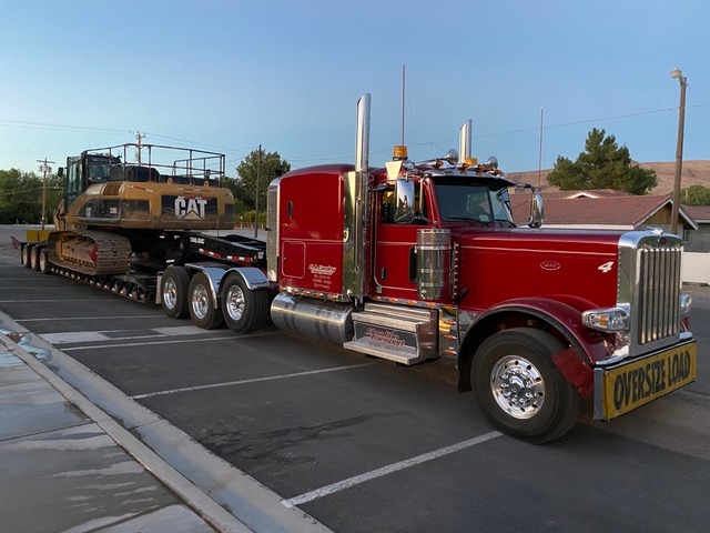 Red truck with a chrome grille towing a large CAT excavator on a flatbed trailer, parked in a lot during twilight.