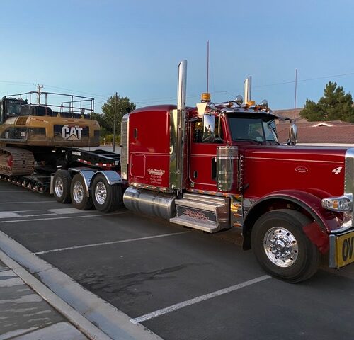 A bright red Peterbilt truck towing a large Caterpillar excavator on a flatbed trailer, parked in a residential area under a clear sky.