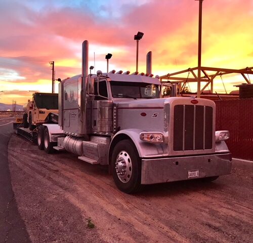 A shiny silver semi-truck with a trailer is parked on a dirt road at sunset, with vibrant clouds in shades of orange and pink in the background.