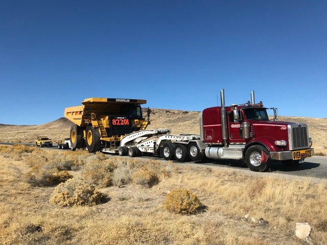 A red semi-truck towing a large yellow Caterpillar dump truck along a rural road, surrounded by dry, hilly terrain under a clear blue sky.