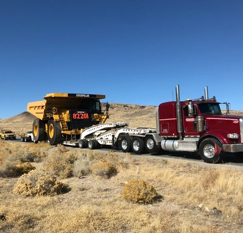 A red truck tows a large yellow Caterpillar dump truck on a flatbed trailer across a dry, hilly landscape under a clear blue sky.
