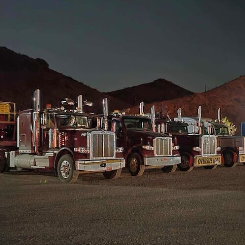 Four large trucks with polished chrome accents are parked under night sky, showcasing heavy-duty vehicles prepared for transport.