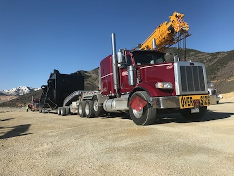 A large red semi-truck with a crane on top tows an oversized black machinery trailer in a mountainous, gravel area.