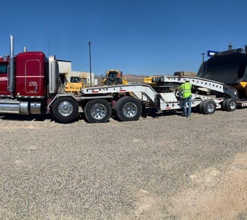 A red semi truck with a lowboy trailer is parked on gravel, while a worker in a reflective vest inspects the equipment. The scene highlights transport and logistics in construction.