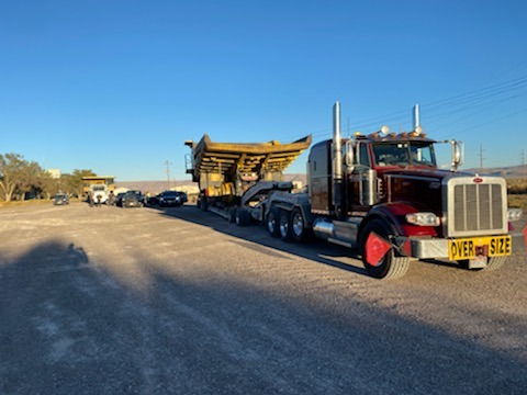 A large red and silver truck hauls an oversized construction vehicle on a gravel road, showcasing its "Over Size Load" sign. The scene illustrates transportation logistics.