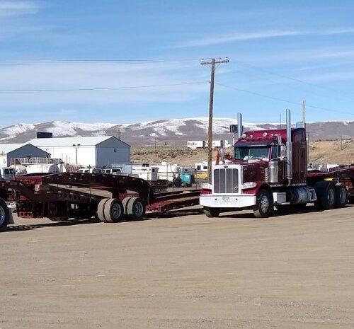 A red and white semi-truck parked next to a flatbed trailer in an industrial lot, with snow-capped mountains in the background.