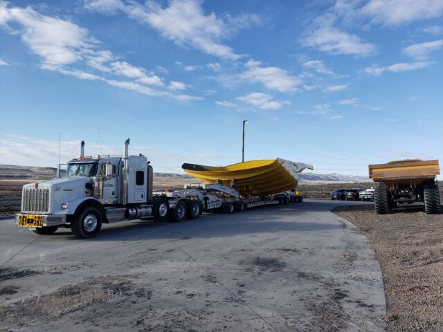 A large yellow construction equipment is being transported by a white semi-truck, surrounded by rugged terrain and blue sky.