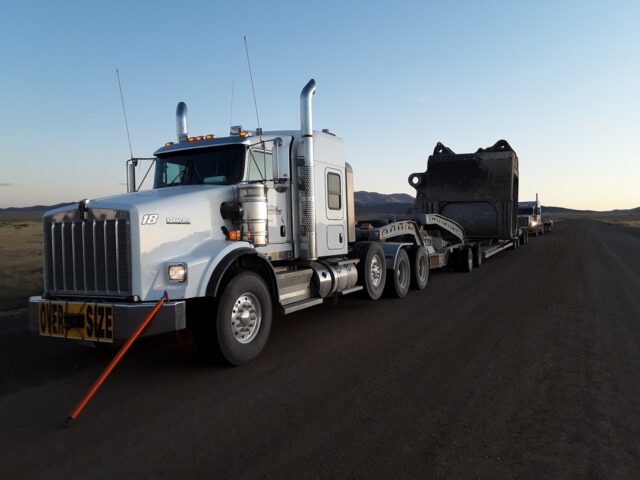 A white semi-truck with oversized load signage is parked on a dirt road, towing a large black piece of equipment against a mountainous backdrop.