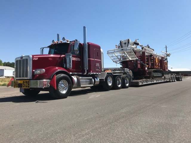 A red semi-truck transports heavy machinery along a road, showcasing its capability to haul large equipment. The clear blue sky highlights the scene.